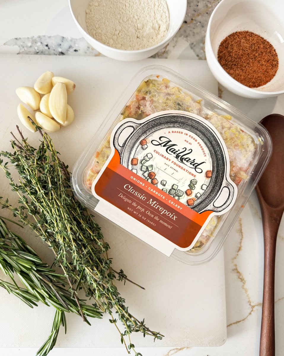 A sealed pack of NO.01 Classic Mirepoix on a cutting board with fresh rosemary, thyme, garlic and a wooden spoon