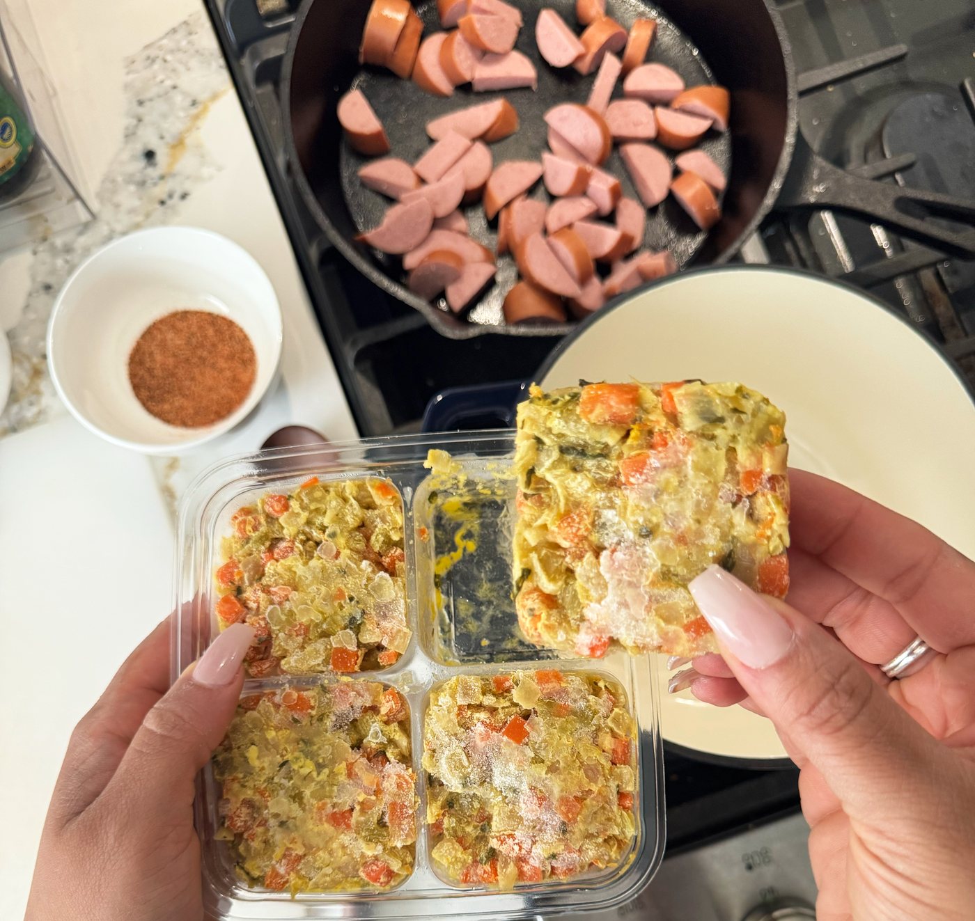 One frozen segment lifted from the pack, with sausage browning in a cast iron skillet behind