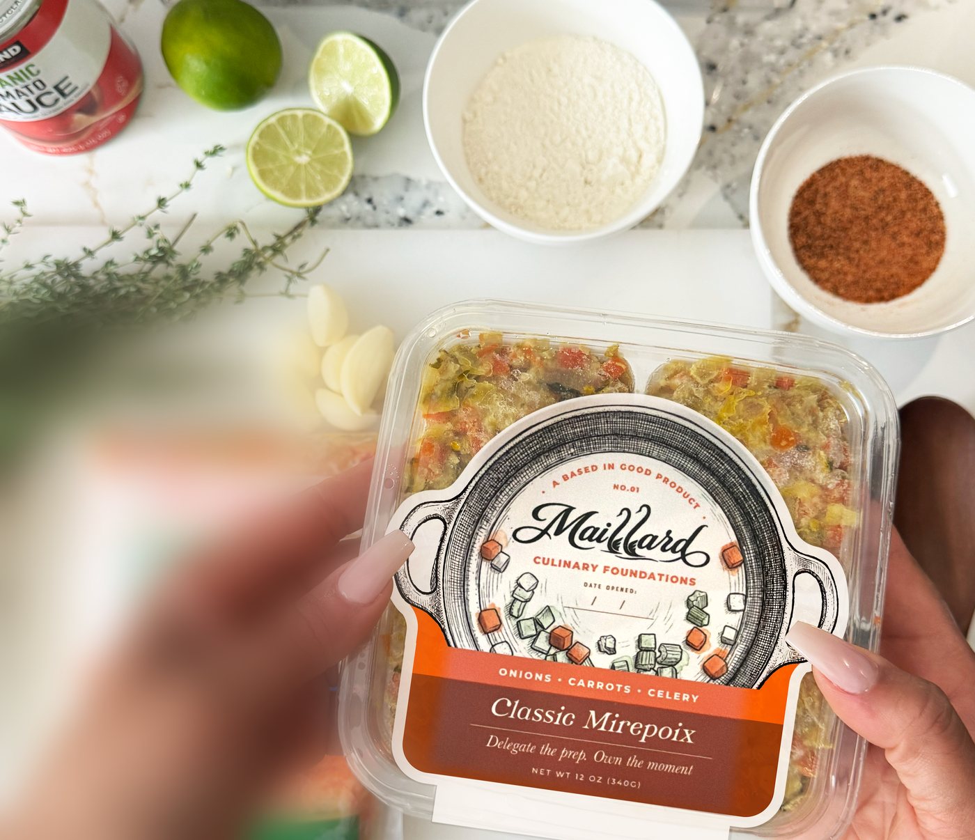 Hands holding a pack of NO.01 Classic Mirepoix above a counter set with limes, garlic, thyme and spices